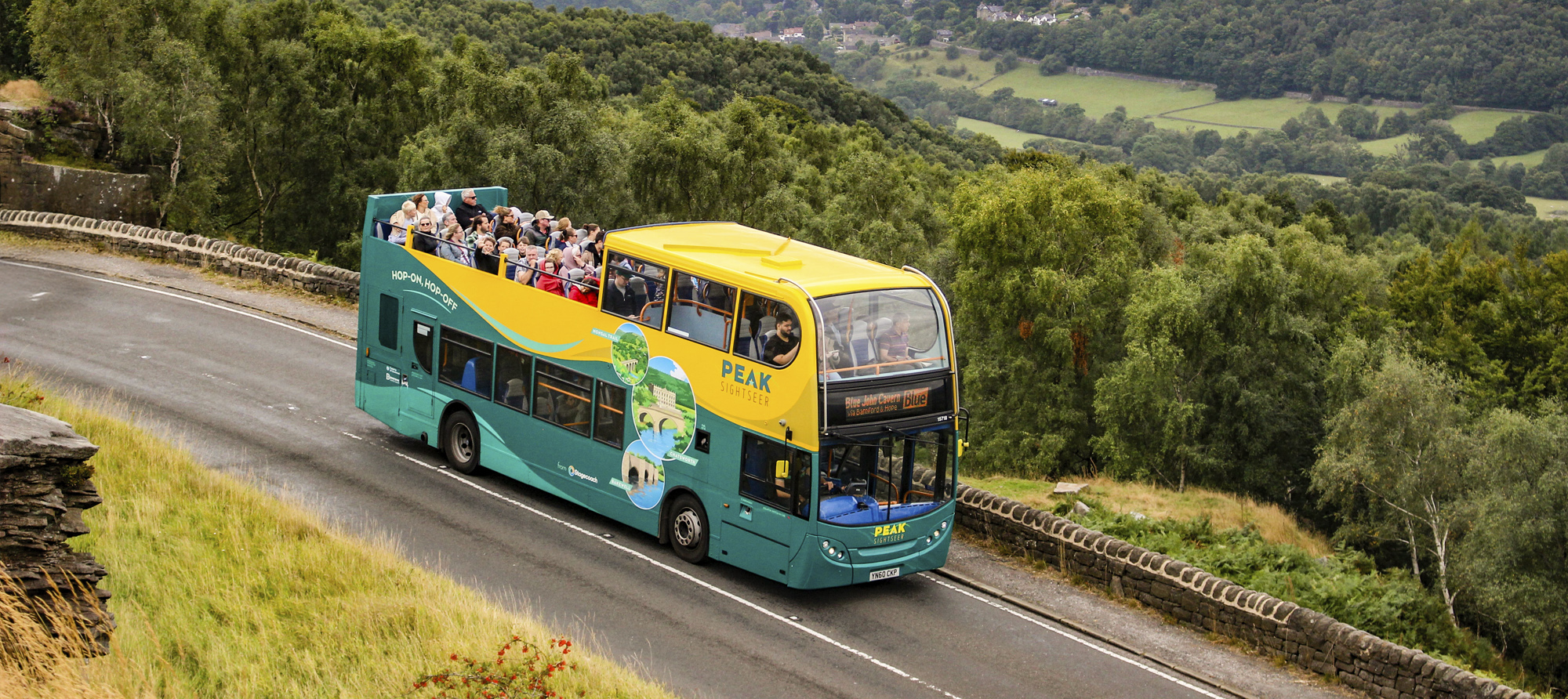 A photograph of a packed Peak Sightseer bus in its stylish wavy teal and yellow livery as it traverses the Peak District countryside
